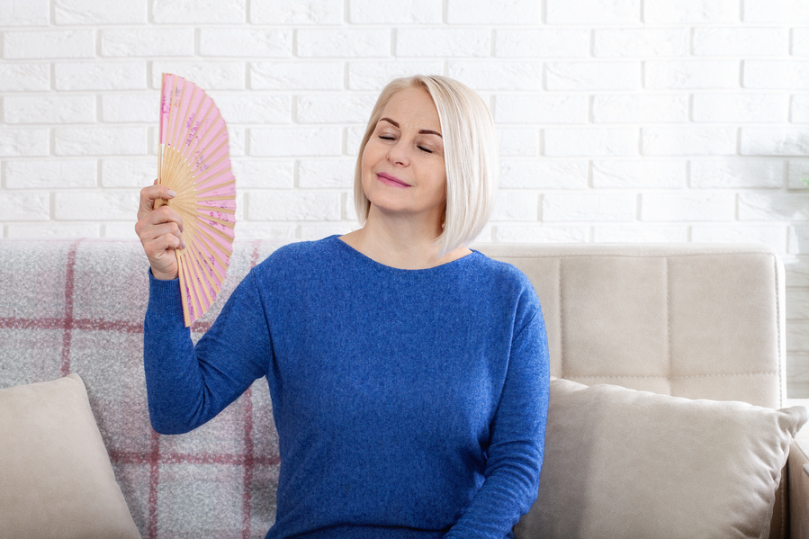 Mature woman experiencing hot flush from menopause. This photo captures the discomfort of hot flashes during menopause, as a woman struggles to cool herself with a delicate paper fan.